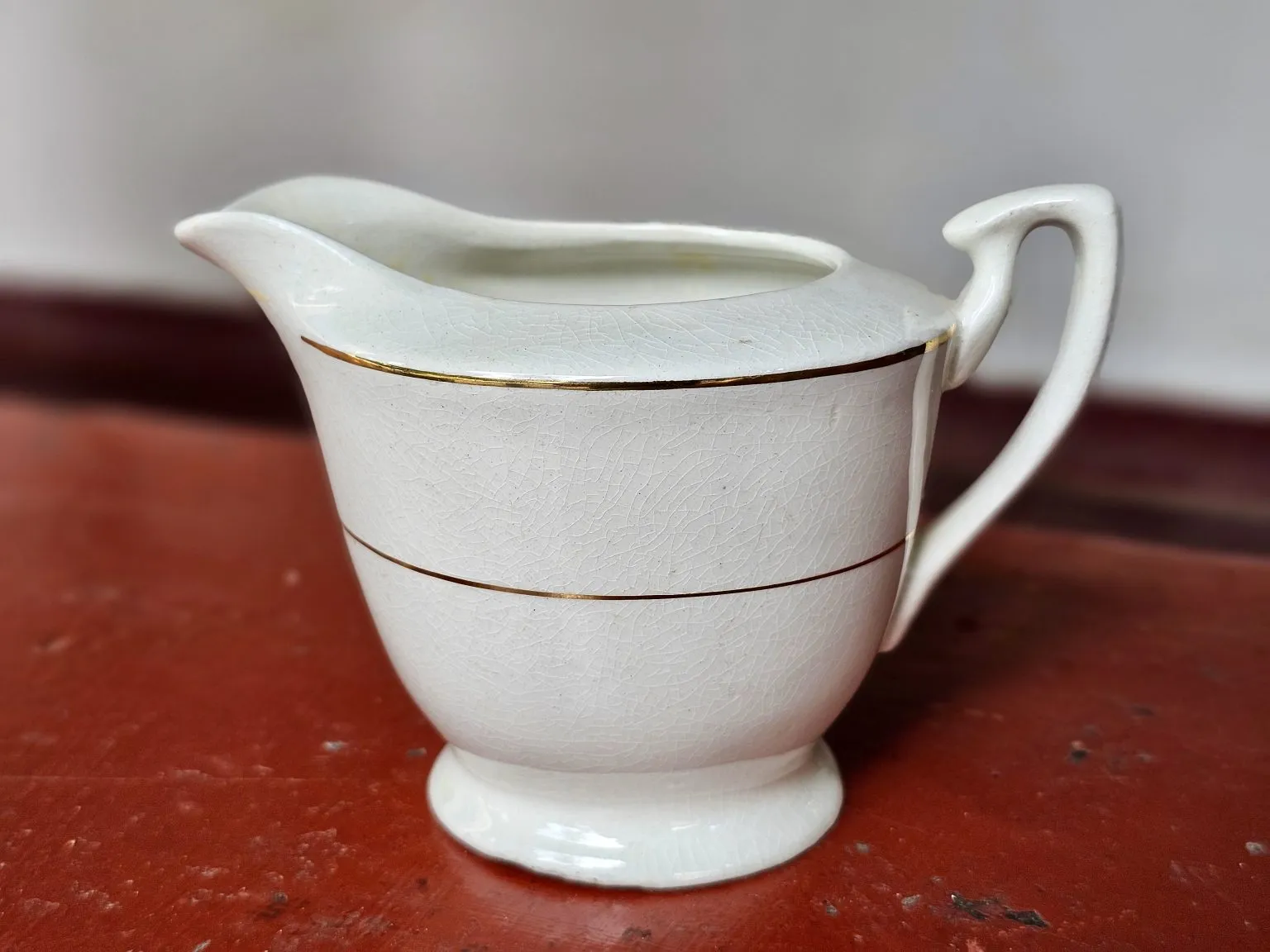 A white ceramic jug with two thin gold bands stands on a red surface.