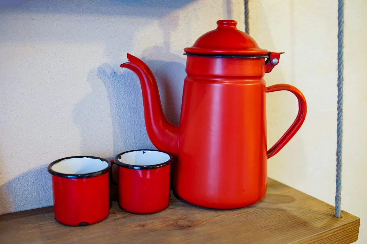 A bright red enamel coffee pot and two matching cups stand on a wooden shelf.